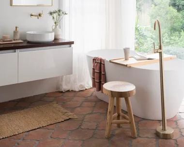 A mediterranean coastal style bathroom with a large white freestanding bath, brass hardware, arched mirror and view on to surrounding bushland.