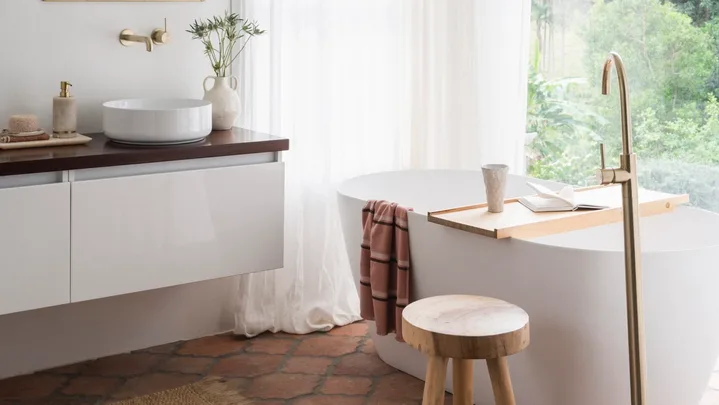 A mediterranean coastal style bathroom with a large white freestanding bath, brass hardware, arched mirror and view on to surrounding bushland.