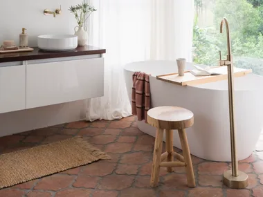 A mediterranean coastal style bathroom with a large white freestanding bath, brass hardware, arched mirror and view on to surrounding bushland.