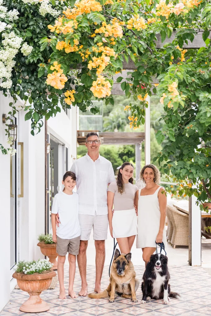 Two parents with two children and two dogs outside their home with yellow bougainvillea flowers
