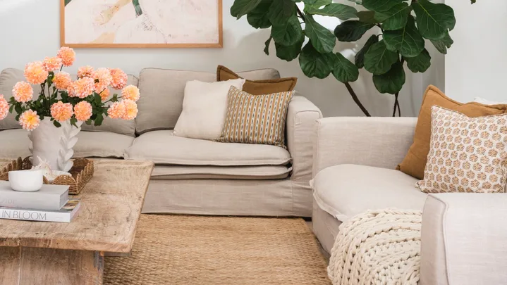 A living room in Mediterranean coastal style with a large plant, a tactile woven rug and cream coloured sofas. A green painting is on the white wall.
