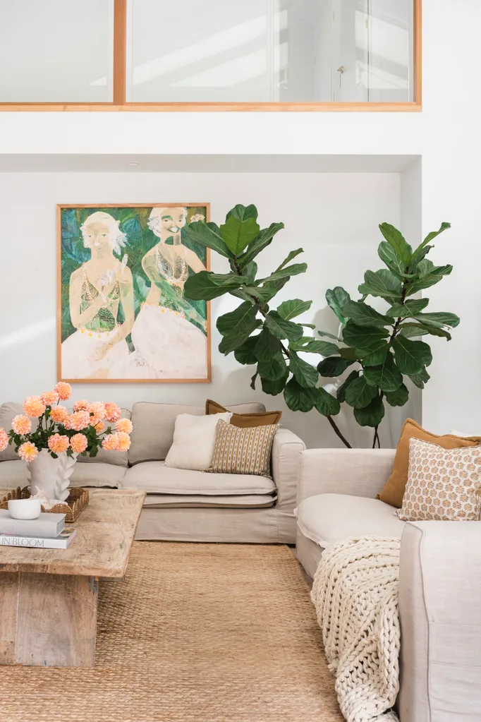 A living room in Mediterranean coastal style with a large plant, a tactile woven rug and cream coloured sofas. A green painting is on the white wall.