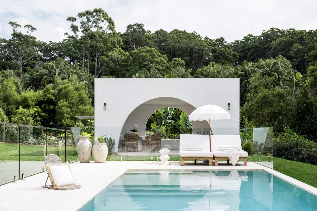 A pool with a white pool room behind it, with a large arch as the entryway. Behind the pool room is luscious bushland.