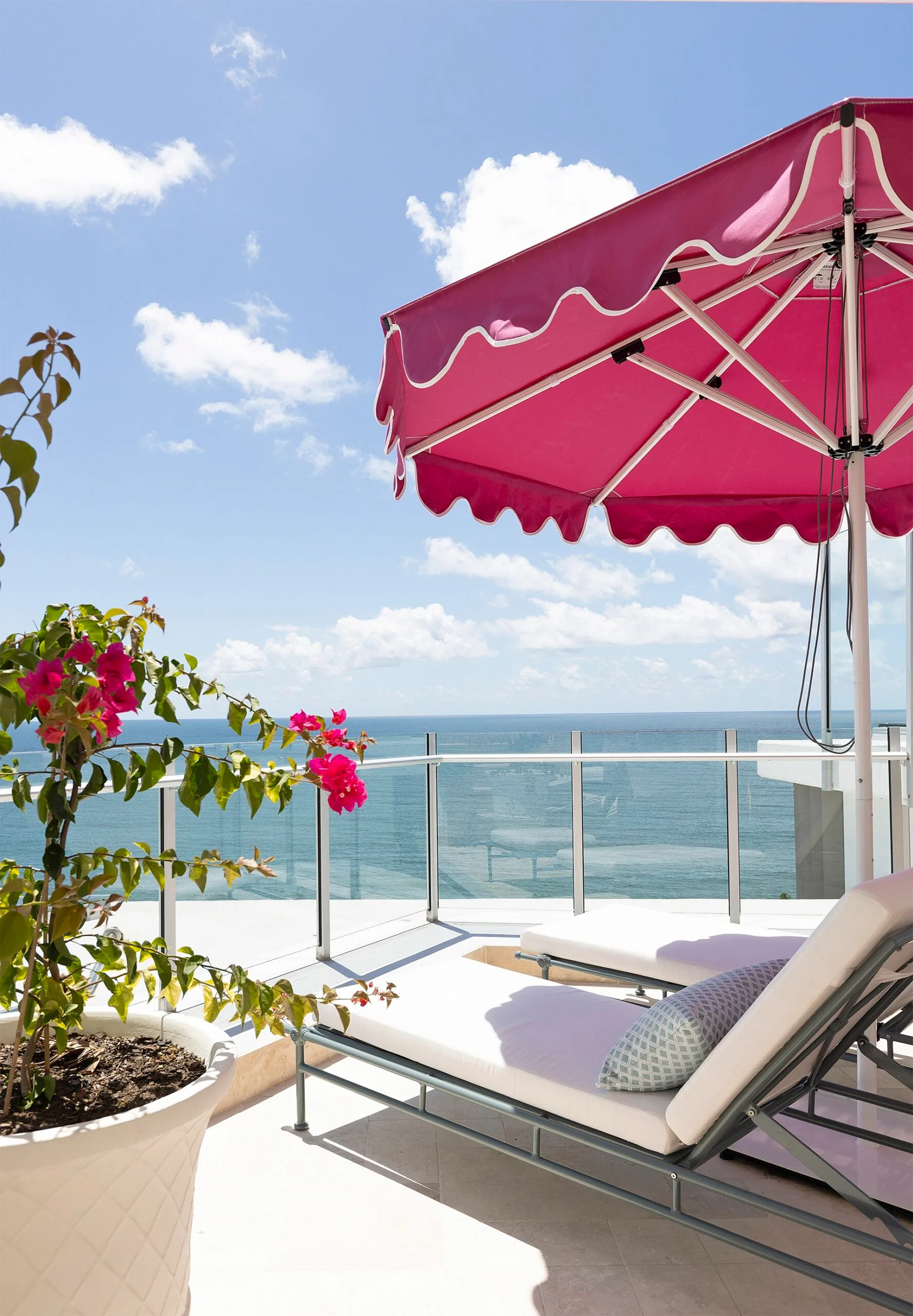 A penthouse apartment balcony with ocean views, a pink beach umbrella and white outdoor lounge and dining furniture.