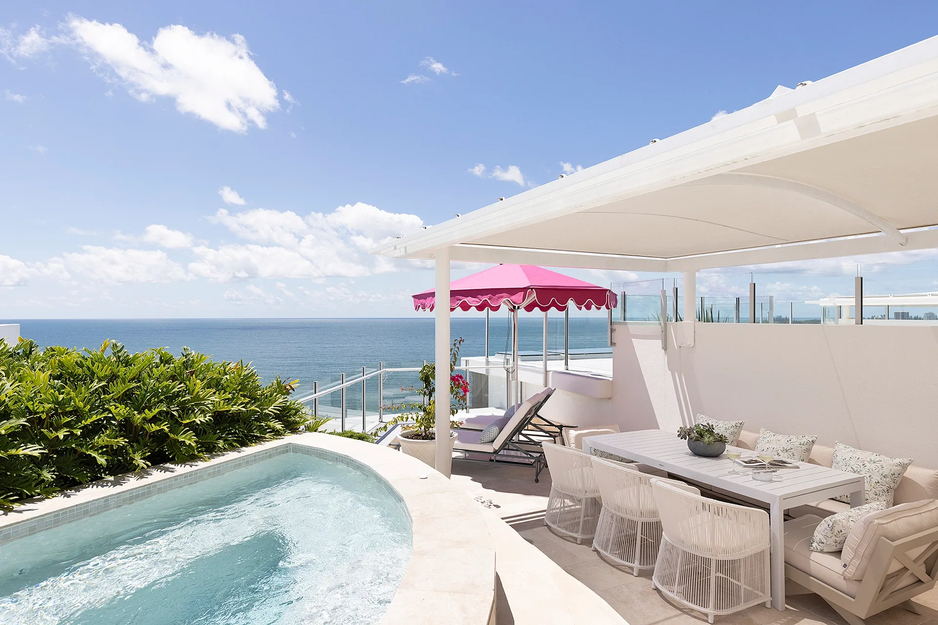 A penthouse apartment balcony with ocean views, a pink beach umbrella and white outdoor lounge and dining furniture.