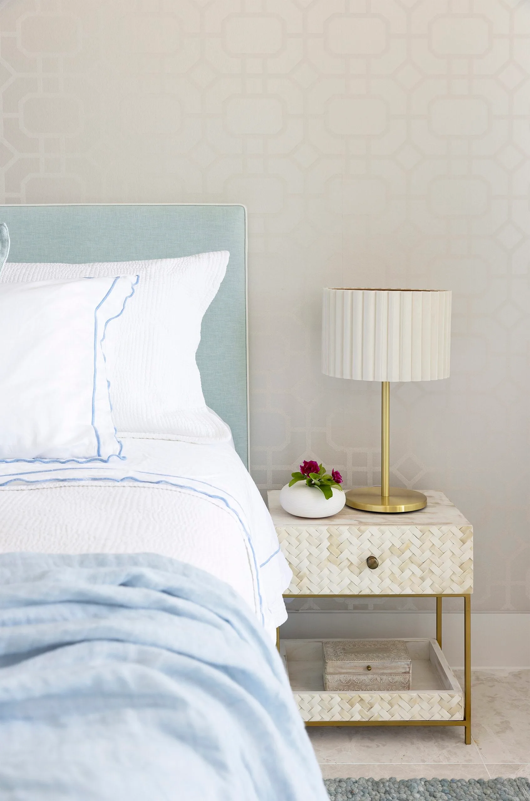 A blue bedhead with white bedding beside a white bedside table with a gold and white lamp on top.