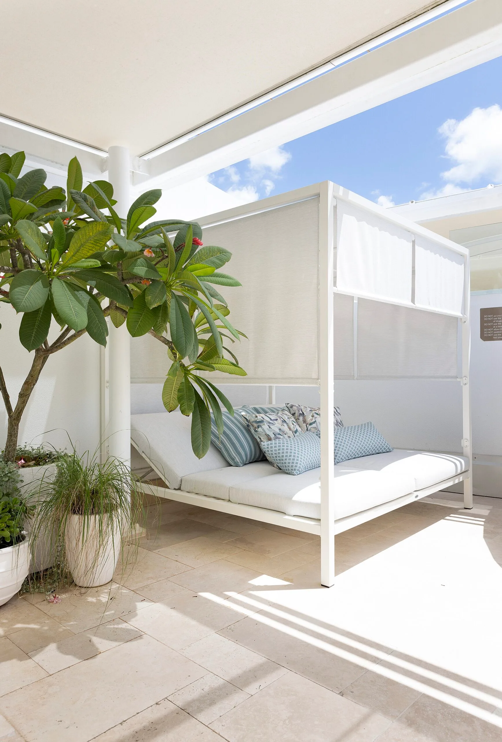 A white daybed with blue cushions in an outdoor area with greenery.