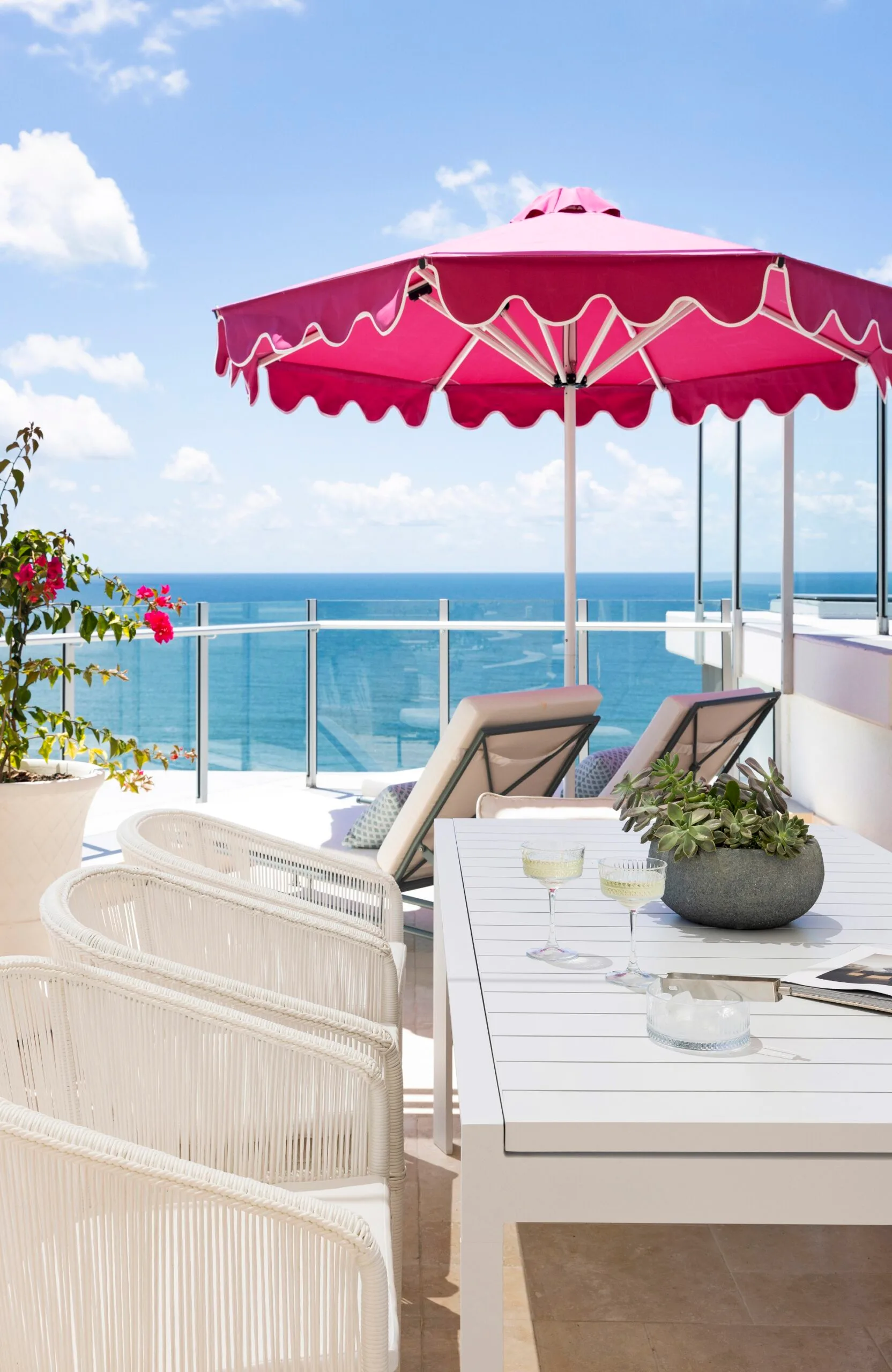 A penthouse apartment balcony with ocean views, a pink beach umbrella and white outdoor lounge and dining furniture.