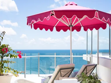 A penthouse apartment balcony with ocean views, a pink beach umbrella and white outdoor lounge and dining furniture.