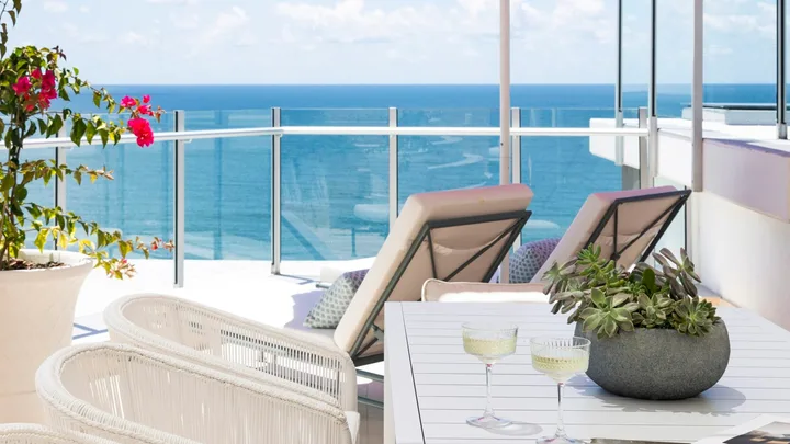 A penthouse apartment balcony with ocean views, a pink beach umbrella and white outdoor lounge and dining furniture.