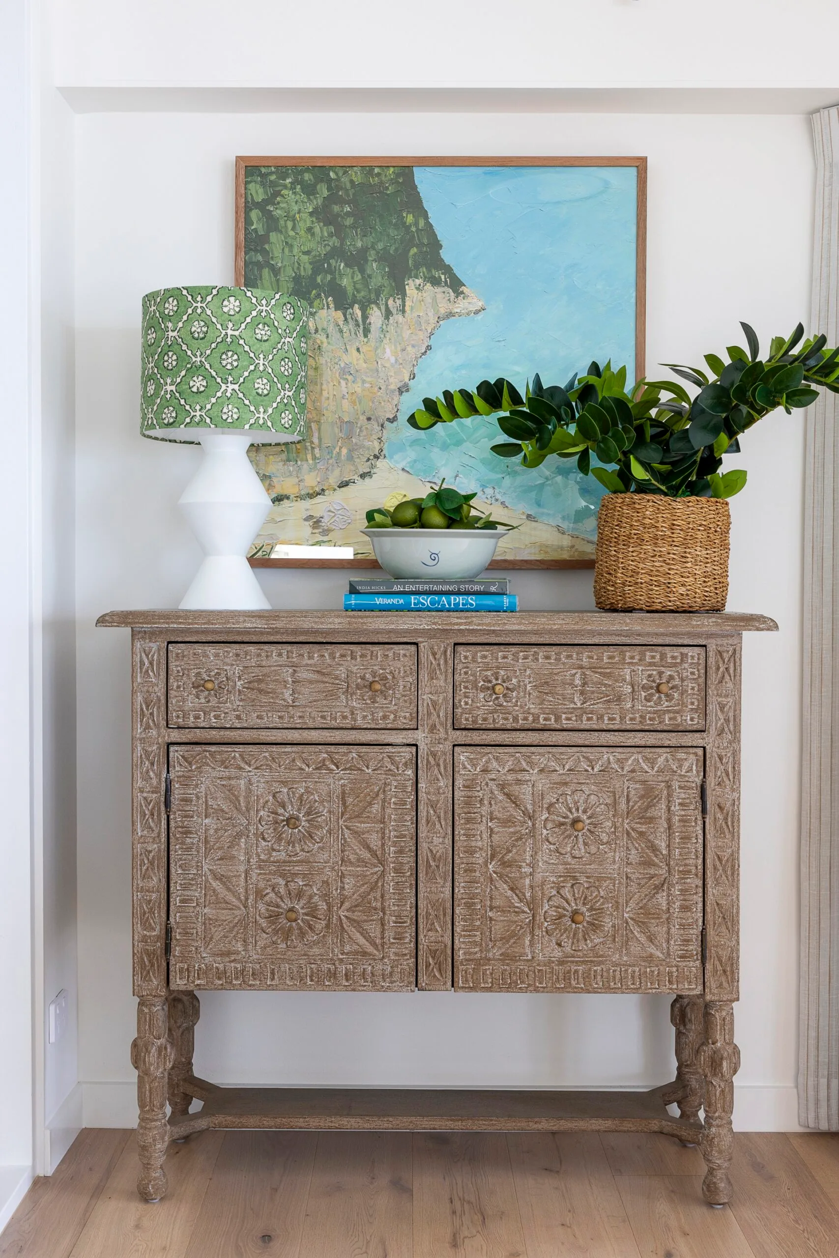 A wooden console table with a painting of a sea cliff, potted plant and a white lamp with a green shade on top.