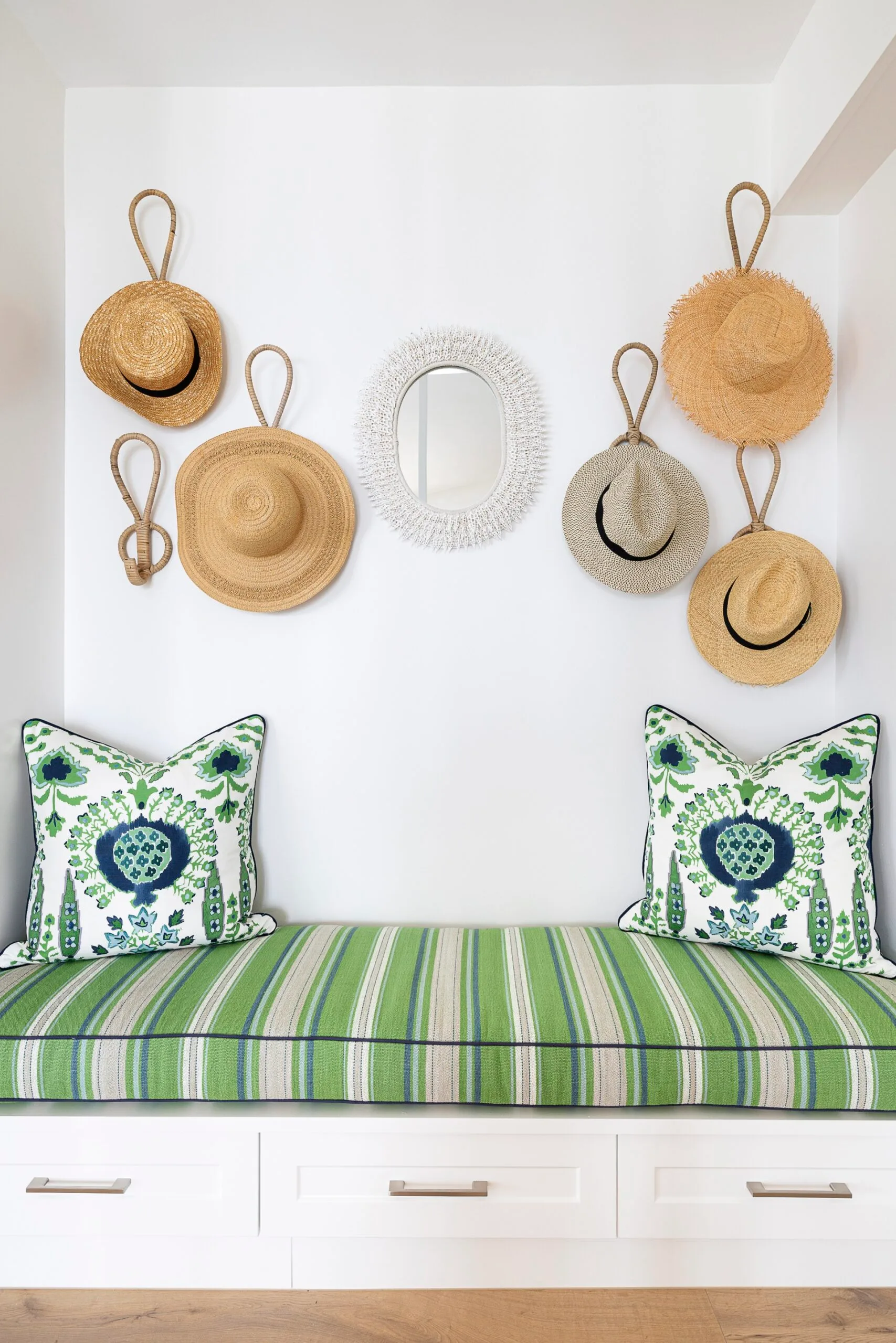 A built in bench with a white base, striped green cushion and woven hats displayed on the wall as decor.