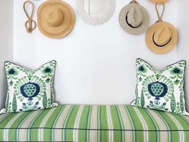 A built in bench with a white base, striped green cushion and woven hats displayed on the wall as decor.