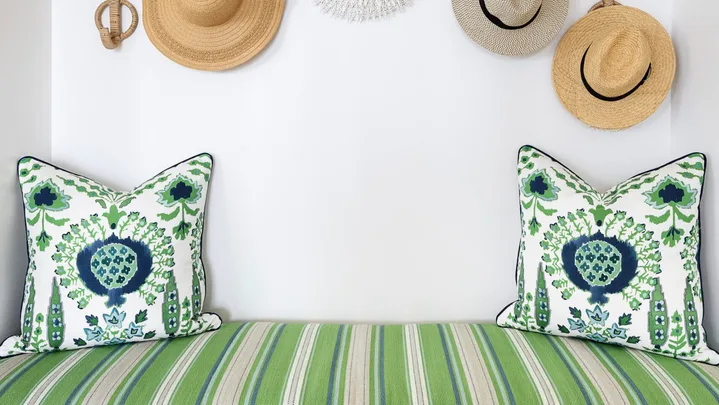 A built in bench with a white base, striped green cushion and woven hats displayed on the wall as decor.