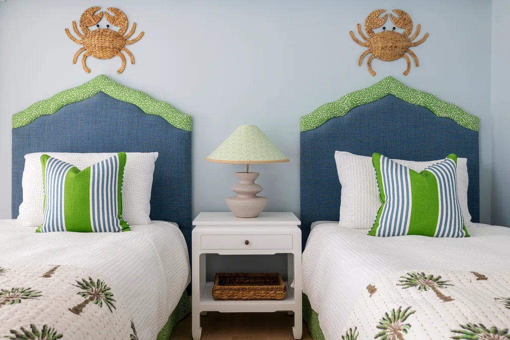 Two single beds with blue and green bedheads, woven crab wall decor and a white bedside table in between the beds.