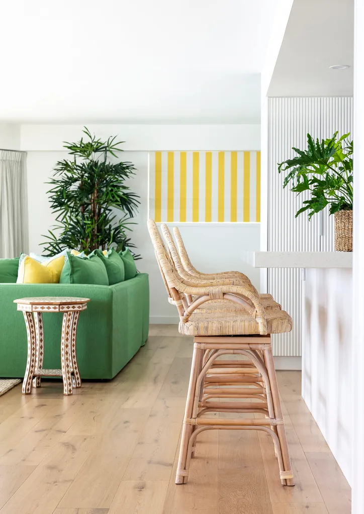 A white kitchen bench with woven bar stools and a living area in the background, with white and yellow striped blinds and a large potted plant.