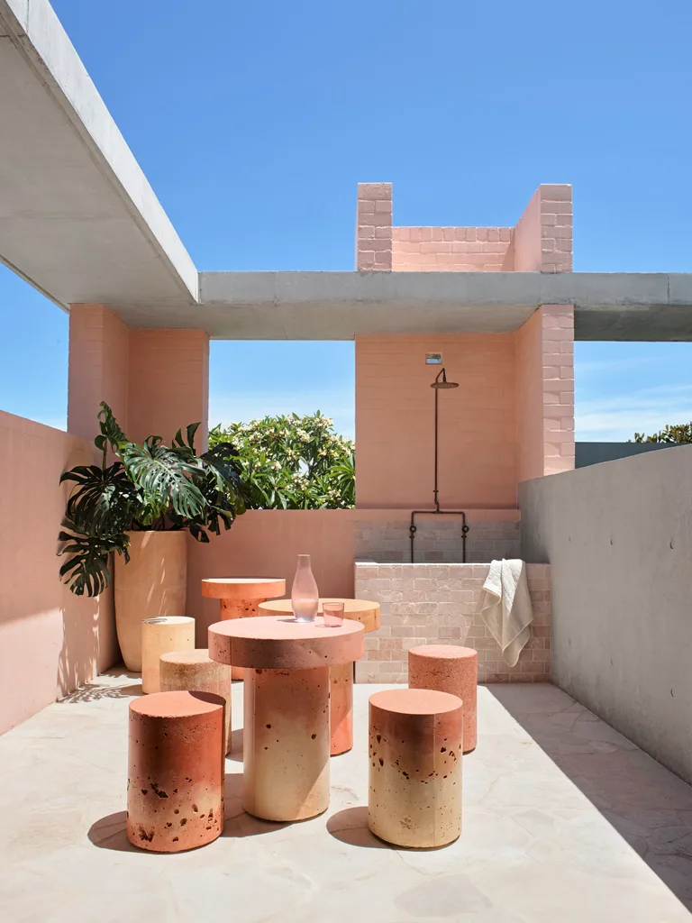 A pink toned outdoor space with circular outdoor stools, table and an outdoor shower in the background.