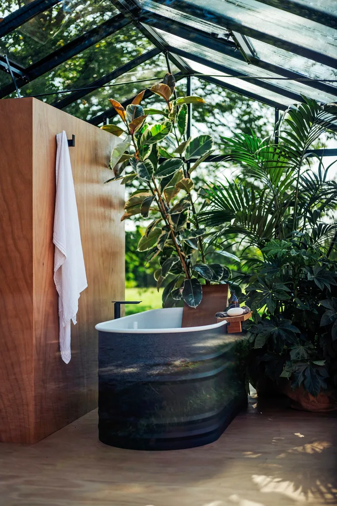 A bath next to a wooden wall with a white towel hanging up, with abundant greenery surrounding.