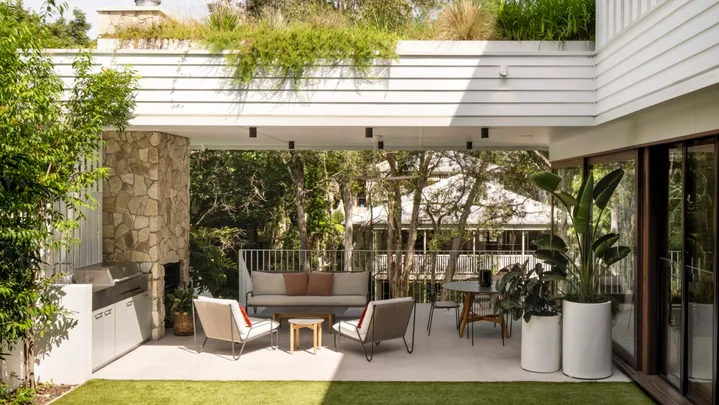 A white exterior of a home. A stone clad fireplace has a chimney that reaches to the white terrace that stretches above the alfresco barbecue kitchen and dining area. There's a strong indoor outdoor connection with alfresco dining and alfresco lounge furniture with a barbecue.