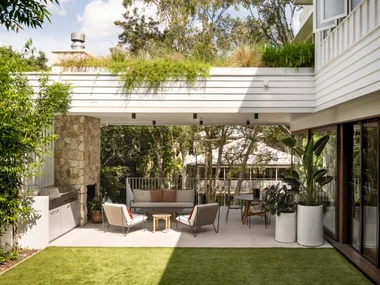 A white exterior of a home. A stone clad fireplace has a chimney that reaches to the white terrace that stretches above the alfresco barbecue kitchen and dining area. There's a strong indoor outdoor connection with alfresco dining and alfresco lounge furniture with a barbecue.