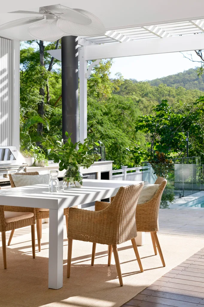 An alfresco dining area with an outdoor cooking zone which has a barbecue, beside a pool. Greenery surrounds the space and tactile woven chairs pair with a white dining table.