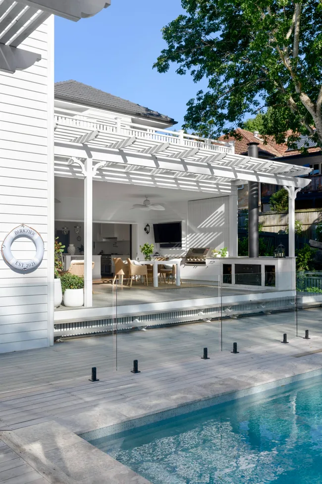 An alfresco dining area with an outdoor cooking zone which has a barbecue, beside a pool. The exterior of the home is white.