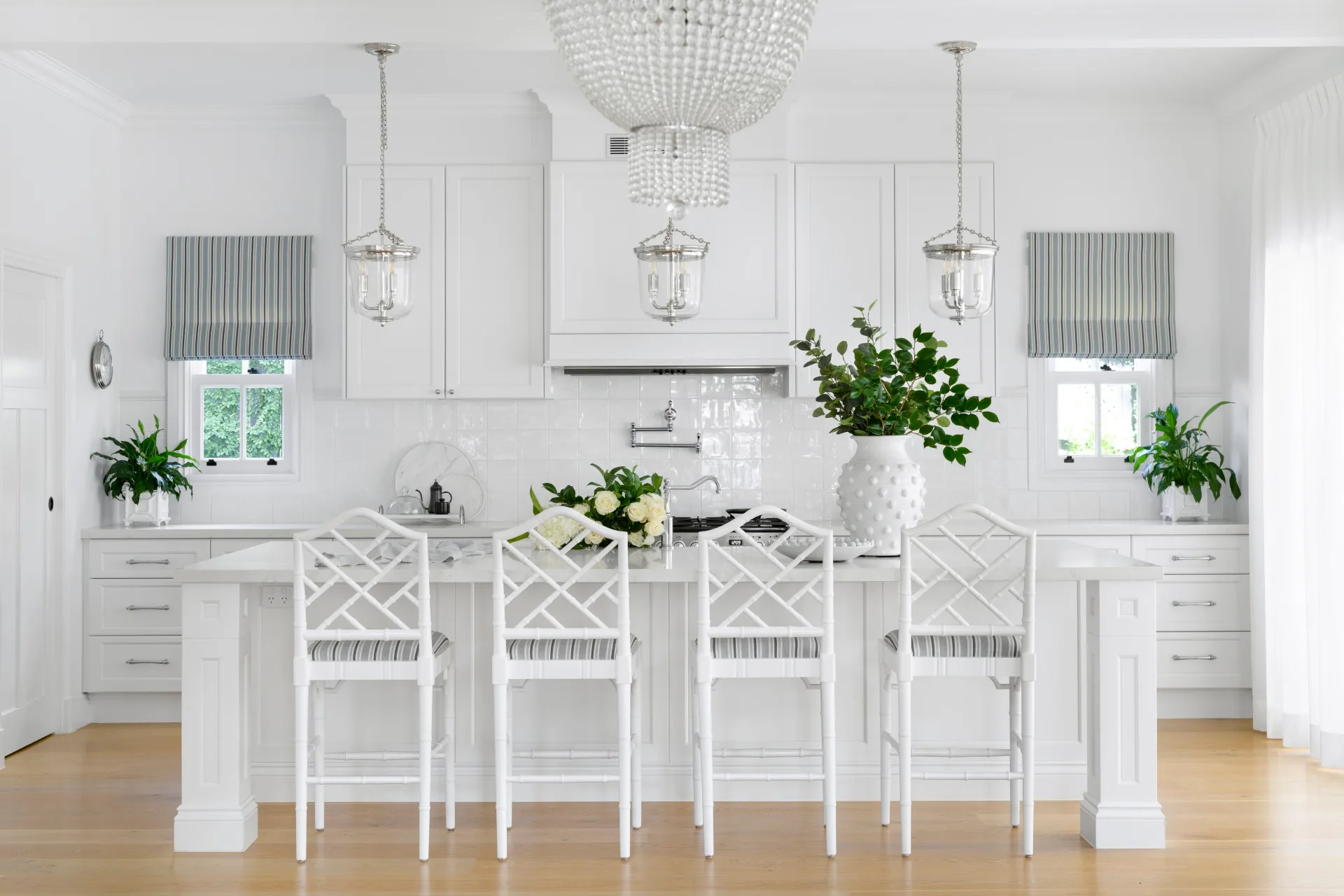 Timber flooring with a white kitchen, which features three pendants above a kitchen island, plus a chandelier. White joinery is beside a white splashback, with a white vase on the kitchen island and white stools around the kitchen island.