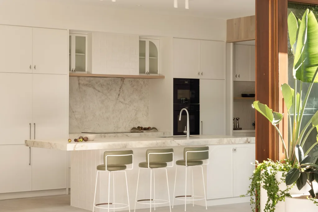 A kitchen with white joinery, a large island and three stools which have white legs and a soft green upholstered seat. The dining area sits beside the kitchen and has expansive corner timber doors that slide back to reveal the outdoor area, tree gully and pool