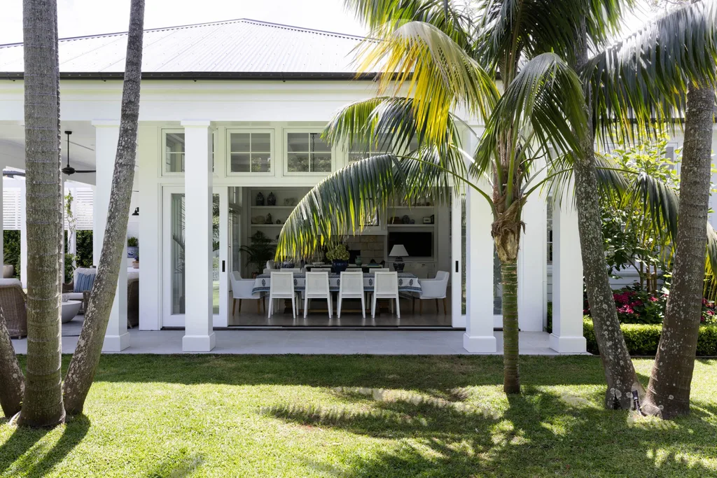 A garden with palm trees and a white exterior. The alfresco dining area has white chairs against a timber floor.