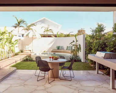 An alfresco dining area with a barbecue with a curved white wall around a circular plunge pool in the background. Green and white striped cushions decorate the bench around the pool and stone is used for the flooring of the outdoor holiday homes dining area.