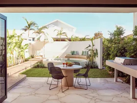 An alfresco dining area with a barbecue with a curved white wall around a circular plunge pool in the background. Green and white striped cushions decorate the bench around the pool and stone is used for the flooring of the outdoor dining area.