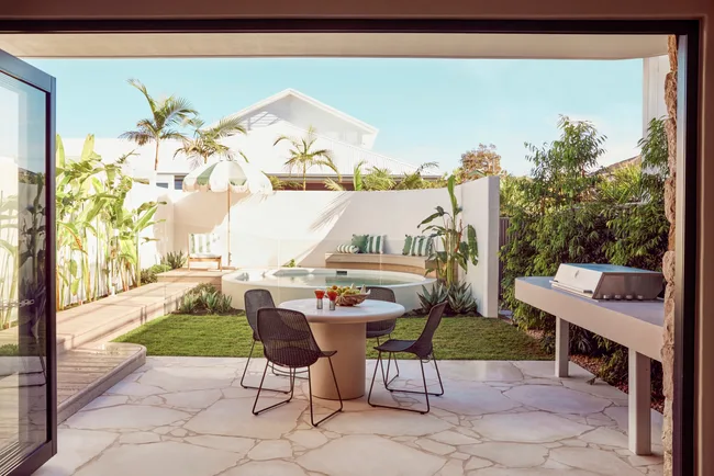 An alfresco dining area with a barbecue with a curved white wall around a circular plunge pool in the background. Green and white striped cushions decorate the bench around the pool and stone is used for the flooring of the outdoor holiday homes dining area.