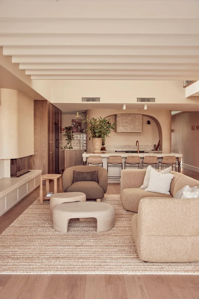 A warm toned open plan kitchen, dining and lounge in a duplex house, with neutral shades and curved furniture, including a curved alcove that fits the stovetop and rangehood in the kitchen.
