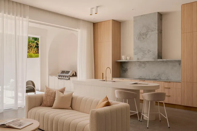 A kitchen designed by Smout Property that has a strong indoor outdoor connection with doors that open fully so that the outdoor barbecue connects to the indoor kitchen, complete with timber joinery, a white curved island and a soft neutral tone curved sofa in the foreground.