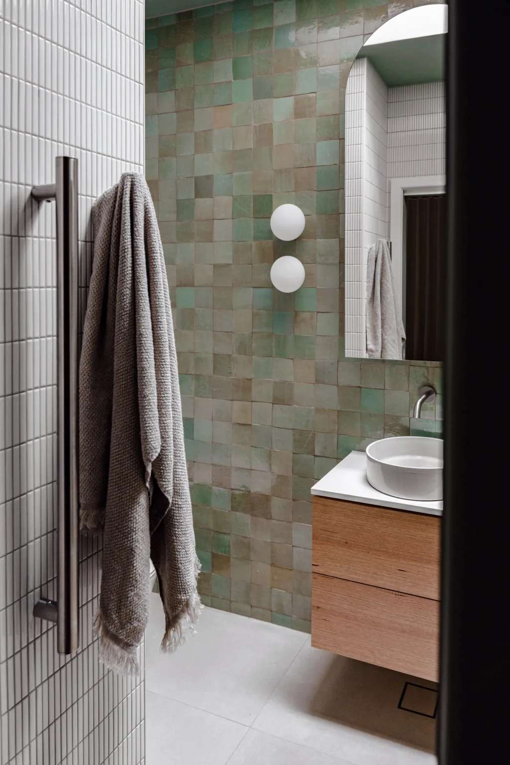 An ensuite bathroom with green zellige handmade wall tiles, two round wall sconces, a curved basin, tapware in brushed nickel, wooden joinery and white kitkat wall tiles.