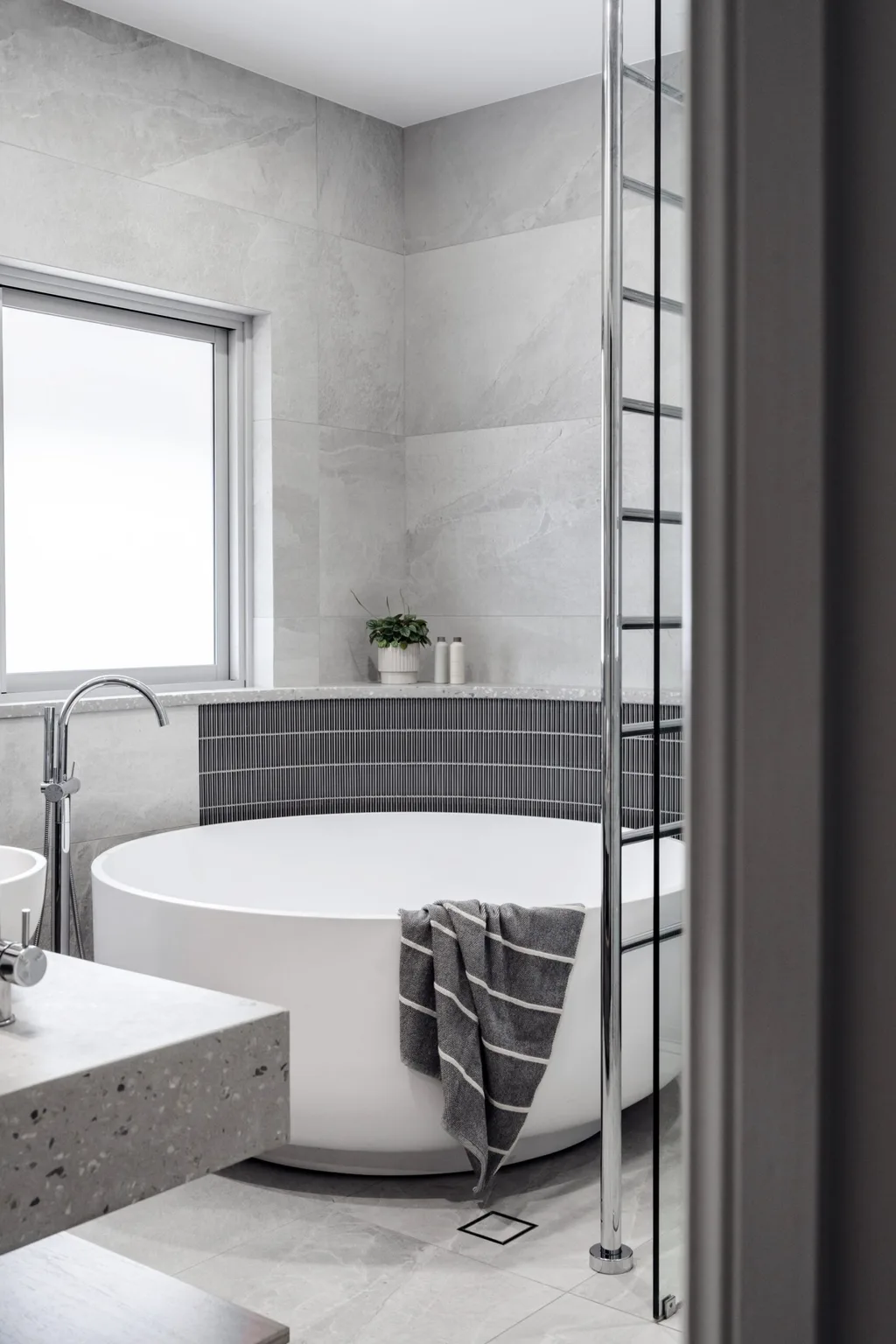 A bathroom shared by two teenage boys which features a round freestanding bath, tapware in a curved style in brushed nickel, grey stone tiles on the wall and floors and grey kitkat tiles on the curved wall around the bath.