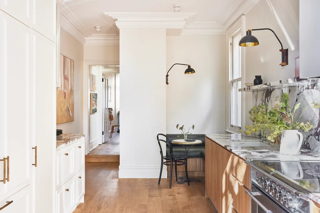 A renovated galley kitchen in a terrace house in Sydney, with a breakfast nook and built-in banquette seating at one end of the kitchen. Marble with burgundy veining was used on the benchtop, rising up to form the splashback then culminating in an open shelf. Timber underbench cabinetry lines one wall while white cabinetry lines the other.