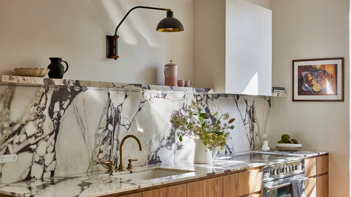 Timber underbench joinery with calacatta viola marble forming the benchtop and splashback. The kitchen set is in Rumbled Brass Organic, with a black wall light positioned above. The rangehood above the oven and stovetop has a white cover which matches the wall beside it.