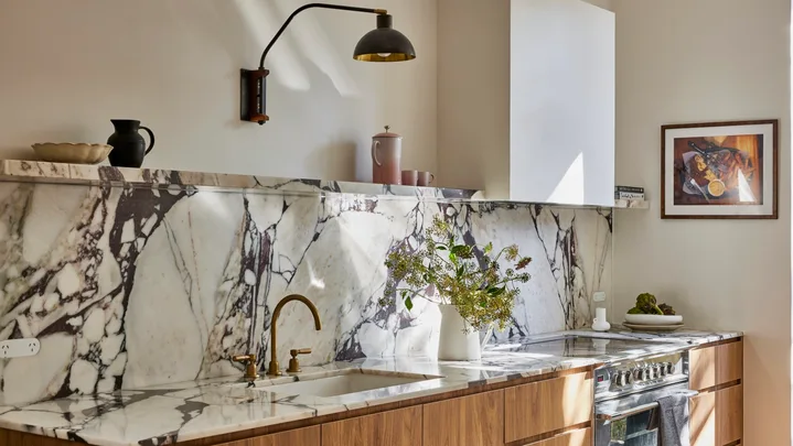 Timber underbench joinery with calacatta viola marble forming the benchtop and splashback. The kitchen set is in Rumbled Brass Organic, with a black wall light positioned above. The rangehood above the oven and stovetop has a white cover which matches the wall beside it.