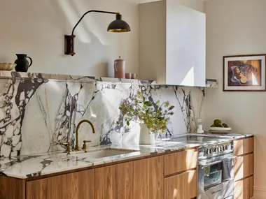 Timber underbench joinery with calacatta viola marble forming the benchtop and splashback. The kitchen set is in Rumbled Brass Organic, with a black wall light positioned above. The rangehood above the oven and stovetop has a white cover which matches the wall beside it.