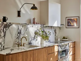Timber underbench joinery with calacatta viola marble forming the benchtop and splashback. The kitchen set is in Rumbled Brass Organic, with a black wall light positioned above. The rangehood above the oven and stovetop has a white cover which matches the wall beside it.