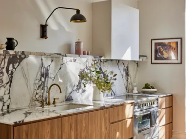 Timber underbench joinery with calacatta viola marble forming the benchtop and splashback. The kitchen set is in Rumbled Brass Organic, with a black wall light positioned above. The rangehood above the oven and stovetop has a white cover which matches the wall beside it.