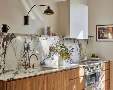Timber underbench joinery with calacatta viola marble forming the benchtop and splashback. The kitchen set is in Rumbled Brass Organic, with a black wall light positioned above. The rangehood above the oven and stovetop has a white cover which matches the wall beside it.