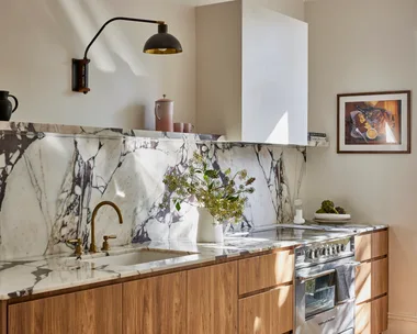 Timber underbench joinery with calacatta viola marble forming the benchtop and splashback. The kitchen set is in Rumbled Brass Organic, with a black wall light positioned above. The rangehood above the oven and stovetop has a white cover which matches the wall beside it.