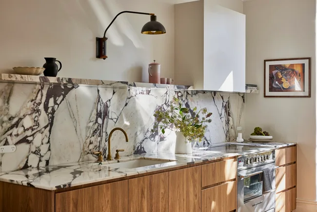 Timber underbench joinery with calacatta viola marble forming the benchtop and splashback. The kitchen set is in Rumbled Brass Organic, with a black wall light positioned above. The rangehood above the oven and stovetop has a white cover which matches the wall beside it.