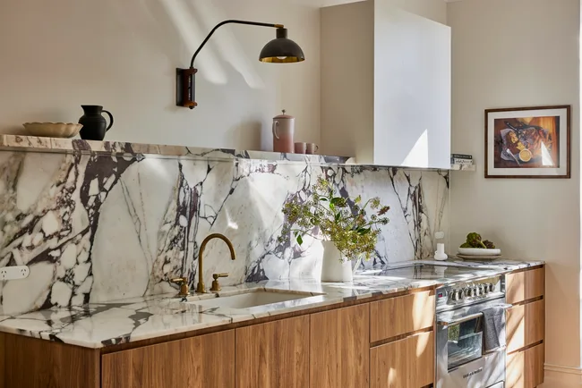 Timber underbench joinery with calacatta viola marble forming the benchtop and splashback. The kitchen set is in Rumbled Brass Organic, with a black wall light positioned above. The rangehood above the oven and stovetop has a white cover which matches the wall beside it.