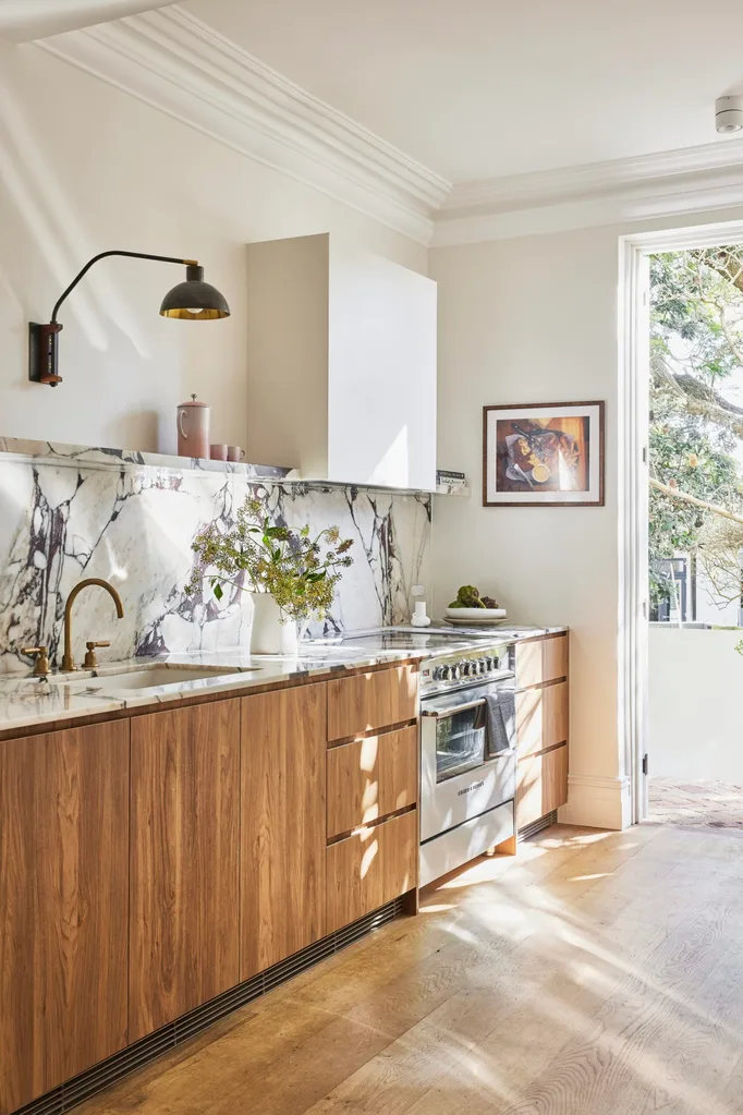 A renovated galley kitchen in a terrace house in Sydney, with French doors at one end of the kitchen. Marble with burgundy veining was used on the benchtop, rising up to form the splashback then culminating in an open shelf. Timber underbench cabinetry lines one wall, with an oven and stovetop placed near the door. Artwork is beside the French doors.
