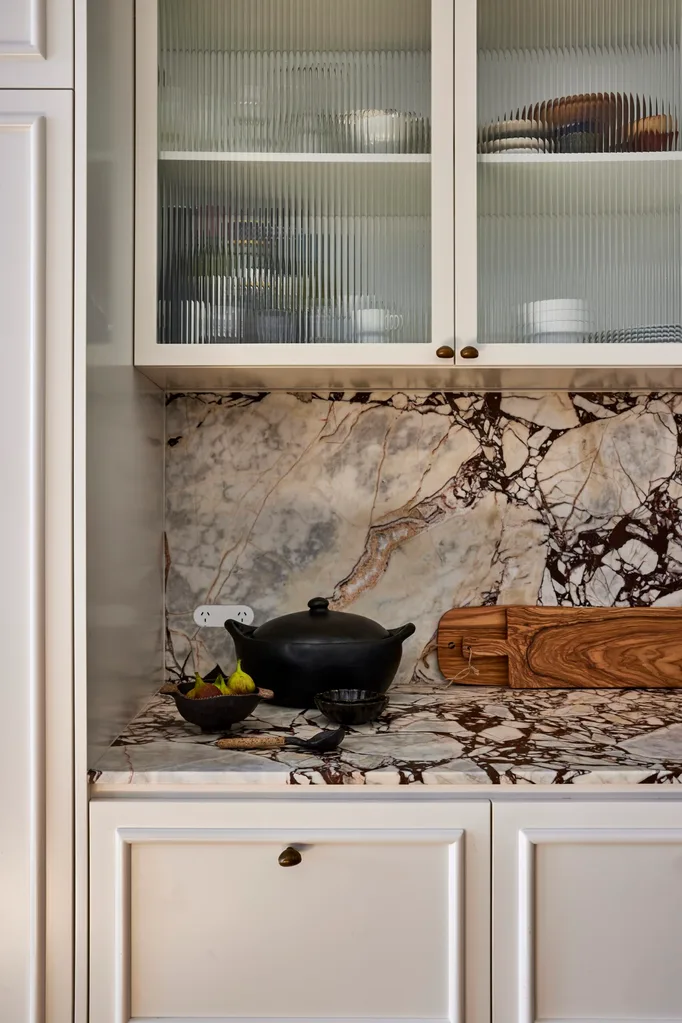 White joinery with bullnose beaded details makes up the underbench cabinetry in this kitchen. Above the marble benchtop and splashback is more cabinetry, with fluted glass fronts.