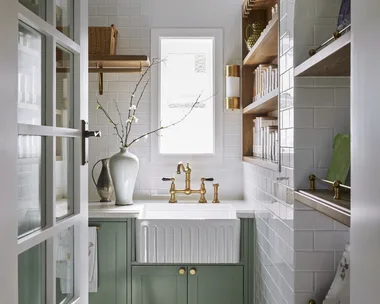 A butler's pantry with white subway tiled walls, a sizable fluted fireclay sink, hardware in Satin Brass and joinery finished in a mint green colour. Timber open shelving allows owners to display trays and dinnerware, plus containers filled with ingredients. The tiled flooring has a star pattern.