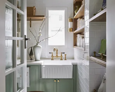 A butler's pantry with white subway tiled walls, a sizable fluted fireclay sink, hardware in Satin Brass and joinery finished in a mint green colour. Timber open shelving allows owners to display trays and dinnerware, plus containers filled with ingredients. The tiled flooring has a star pattern.